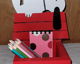 May include: A red wooden desk organizer shaped like Snoopy's doghouse. The white Snoopy figure is on top. Inside are colored pencils and a pink notepad with brown polka dots. A charming and functional desk accessory.