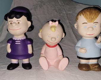 May include: Three ceramic figurines of cartoon characters. The first is a girl in a purple dress and black shoes. The second is a baby in a pink outfit. The third is a boy in a blue shirt and brown shoes.