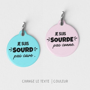 May include: Two round pet tags with silver clasps. One is light blue with the text "JE SUIS SOURD pas cave." in black. The other is pink with the text "JE SUIS SOURDE pas conne." in black. The tags have a circular shape.