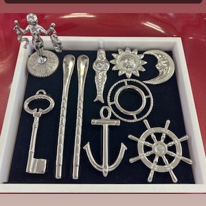 Op de afbeelding: Een verzameling religieuze items van roestvrij staal, waaronder een sleutel, lepels, een anker, een scheepsrad en andere decoratieve figuren. De tekst bovenaan luidt "Herramientas para Santos Acero inoxidable/Stainless Steel". Het woord "Olokun" staat onderaan.