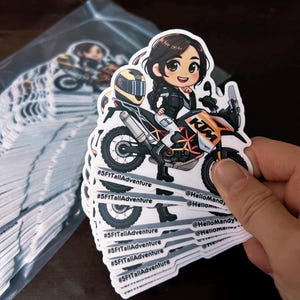 Puede incluir: Una pila de pegatinas blancas con una ilustraci&oacute;n de dibujos animados de una mujer conduciendo una motocicleta. La motocicleta es naranja y blanca con la palabra "KTM" en el lateral. La mujer lleva una chaqueta negra y un casco. Las pegatinas tienen el texto "#5FtTallAdventure" y "@HelloMandy" en ellas.