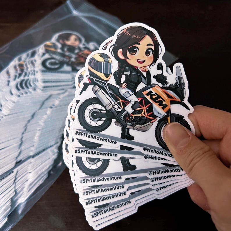 Puede incluir: Una pila de pegatinas blancas con una ilustraci&oacute;n de dibujos animados de una mujer conduciendo una motocicleta. La motocicleta es naranja y blanca con la palabra "KTM" en el lateral. La mujer lleva una chaqueta negra y un casco. Las pegatinas tienen el texto "#5FtTallAdventure" y "@HelloMandy" en ellas.