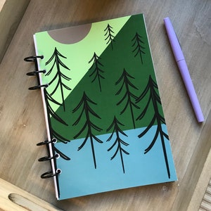 May include: A spiral bound notebook with a green, blue, and brown cover featuring a stylized illustration of a forest with trees and a sun. A purple pen is lying on a wooden tray next to the notebook.