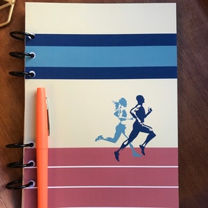May include: Spiral-bound notebook with a running theme. The cover features a runner silhouette in blue and a track design. An orange pen rests beside the notebook. The notebook has a cream-colored background with blue stripes.