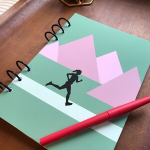 May include: A spiral-bound notebook with a green and pink geometric design featuring a black silhouette of a runner. A red pen rests on the notebook. The notebook is on a wooden tray with a gold candle holder.