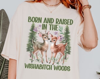 May include: A cream-colored t-shirt with the text "Born and Raised in the Wishabitch Woods" and a graphic of two deer in a forest setting.