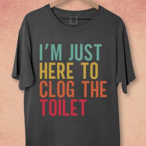 May include: A dark gray t-shirt with the text "I'M JUST HERE TO CLOG THE TOILET" in a rainbow color scheme.