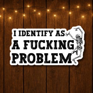 May include: A white sticker with black text that reads "I IDENTIFY AS A FUCKING PROBLEM." A skeleton illustration is on the right side of the text. The sticker is on a wooden background with string lights.