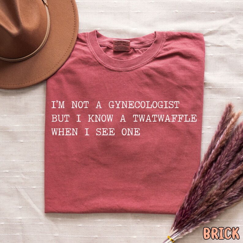 May include: A red t-shirt with the text "I'm not a gynecologist but I know a twatwaffle when I see one" printed in white.