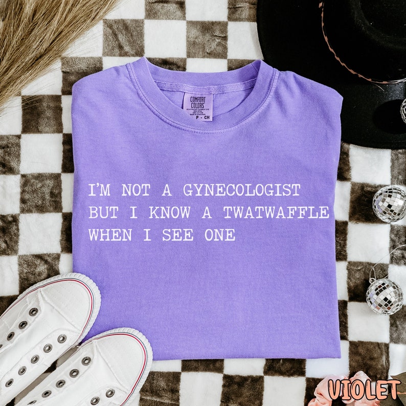 May include: Purple t-shirt with the text "I'm not a gynecologist but I know a twatwaffle when I see one" printed in white.