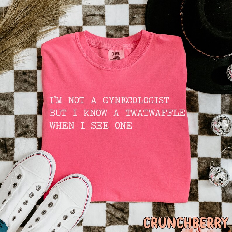 May include: Pink t-shirt with the text "I'm not a gynecologist but I know a twatwaffle when I see one" printed in white.