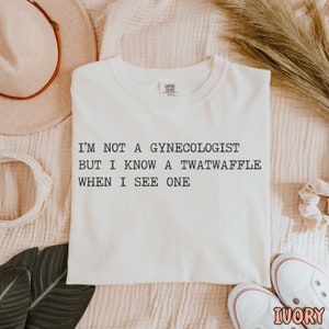 May include: A white t-shirt with the text "I'm not a gynecologist but I know a twatwaffle when I see one" printed in black.