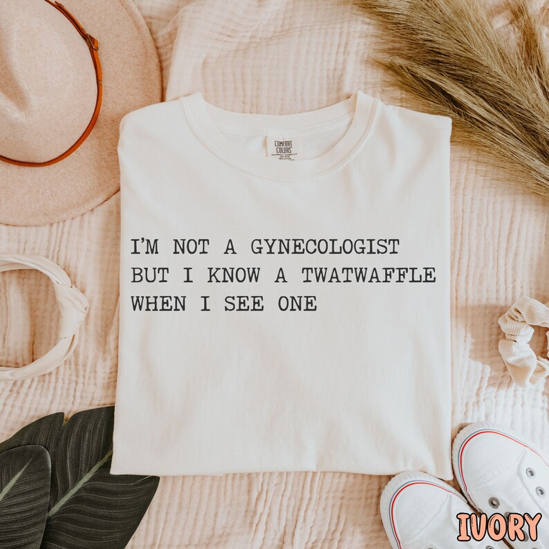May include: A white t-shirt with the text "I'm not a gynecologist but I know a twatwaffle when I see one" printed in black.