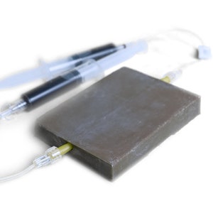 Könnte beinhalten: Zwei klare Spritzen mit schwarzer Flüssigkeit, die mit einem braunen rechteckigen Block und einem durchsichtigen Schlauch verbunden sind.