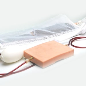 Könnte beinhalten: Ein medizinisches Gerät mit einer weißen Tasche, einem durchsichtigen Schlauch, einem roten Schlauch, einer weißen Birne und einem rechteckigen beigefarbenen Bauteil.