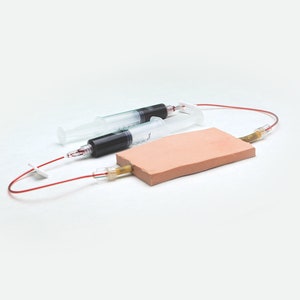 Könnte beinhalten: Zwei klare Spritzen mit schwarzer Flüssigkeit, die mit einem roten Schlauch und einem braunen schwammartigen Pad verbunden sind.