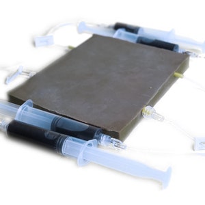 Könnte beinhalten: Nahaufnahme eines grauen rechteckigen Objekts mit vier daran befestigten durchsichtigen Schläuchen. Zwei durchsichtige Spritzen mit schwarzer Flüssigkeit sind an die Schläuche angeschlossen.