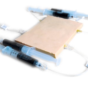 Könnte beinhalten: Drei transparente Kunststoffspritzen mit schwarzer Flüssigkeit sind mit einem beigen medizinischen Übungspad verbunden. Die Spritzen sind an transparente Kunststoffschläuche angeschlossen.