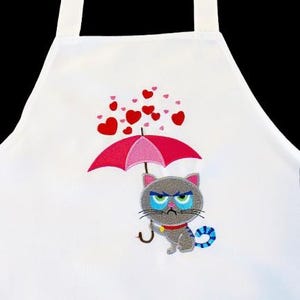 Peut inclure: Tablier blanc avec une broderie d'un chat gris grincheux sous un parapluie rose, avec des cœurs rouges. Le chat a des yeux bleus et verts, un collier rouge et un jouet rayé bleu et blanc. Le tablier a une sangle de cou blanche.