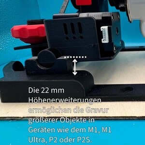Pode incluir: Uma impressora 3D preta com uma ma&ccedil;aneta vermelha e uma extens&atilde;o de altura de 22 mm. O texto "Die 22 mm H&ouml;henerweiterungen erm&ouml;glichen die Gravur gr&ouml;&szlig;erer Objekte in Ger&auml;ten wie dem M1, M1 Ultra, P2 oder P2S." &eacute; vis&iacute;vel na base da impressora.