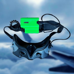 Könnte beinhalten: Ein schwarzes Headset mit einer grünen Box, die an der Kopfband befestigt ist. Das Headset hat zwei Linsen und ein Band, das über den Kopf geht. Die grüne Box hat ein schwarzes Kabel, das daran befestigt ist.