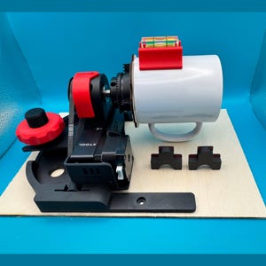 Op de afbeelding: Een zwarte en rode mokpers met een witte mok eraan bevestigd. De mokpers heeft een waterpas aan de bovenkant en een rode knop aan de zijkant. Er liggen twee zwarte rubberen pads op de tafel.