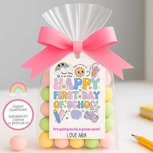 以下が含まれることがあります： ピンクのリボンで結ばれた透明な袋に、カラフルなキャンディーが入っています。「HAPPY FIRST DAY OF SCHOOL」と書かれたタグには、虹、太陽、学校用品のイラストが描かれています。テキストには「It's going to be a great year! LOVE ARIA.」と書かれています。