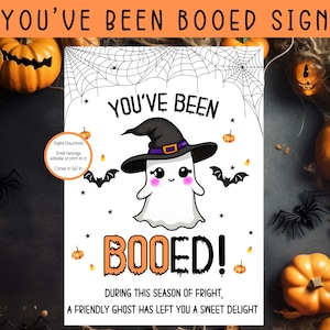 May include: Printable "You've Been Booed" sign featuring a cute ghost wearing a witch hat and holding a candy corn. The sign has a black and white spiderweb background with orange and black text.