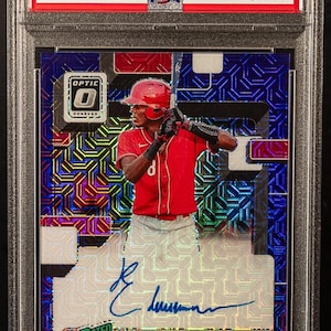 Puede incluir: Tarjeta de béisbol con Elly De La Cruz con uniforme rojo, sosteniendo un bate. La tarjeta tiene un fondo holográfico con el texto "2022 Donruss Optic" y "Rated Prospect". La tarjeta está calificada y encapsulada.