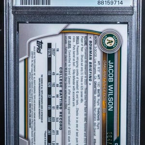 May include: A baseball card encased in a protective holder, featuring Jacob Wilson of the Oakland Athletics. The card displays player stats, team logo, and a PSA authentication label. The card is numbered 064/150.