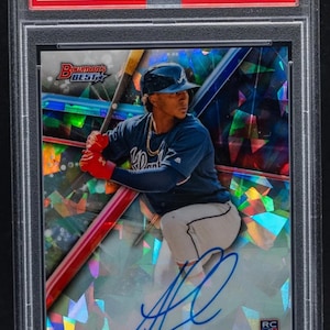 Peut inclure: Carte de baseball d'Ozzie Albies en uniforme bleu des Braves d'Atlanta, dans un étui. La carte est de 2018 Bowman's Best, AU-Atomic, avec un autographe. Numérotée 22/25. La carte est dans un étui de protection.