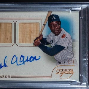 Può includere: Una carta di baseball di Hank Aaron della serie Topps Dynasty 2014. La carta presenta Hank Aaron con una mazza e la sua firma blu. Include due reliquie in legno. La carta è valutata GEM MT 10.