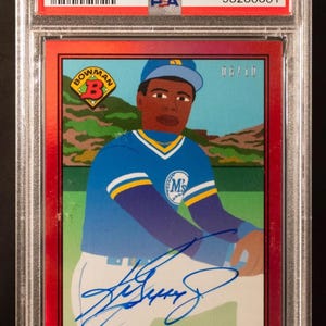 Könnte beinhalten: Eine rot umrandete Sammelkarte mit Ken Griffey Jr. in einer blauen Baseballuniform mit Autogramm. Die Karte ist bewertet und in einem Schutzhalter eingeschlossen. Der Text enthält "2021 Bowman X K.Shore" und "Autograph-Red Foil."