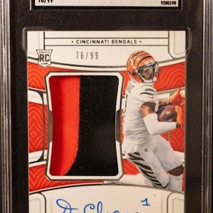 Könnte beinhalten: Eine bewertete Sammelkarte mit Ja'Marr Chase von den Cincinnati Bengals. Die Karte ist eine 2021 Panini National Treasures Rookie Patch Autographed Karte, nummeriert 76/99, mit einer Bewertung von 9,5. Die Karte hat einen schwarzen und roten Patch und den Text "CHASER!!".