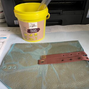 Puede incluir: Un cubo amarillo con un tubo de plástico transparente en su interior, junto a un trozo de cuero y una correa marrón. El cubo tiene una etiqueta con el texto "Klassische Lederpflege". Una impresora está en el fondo.