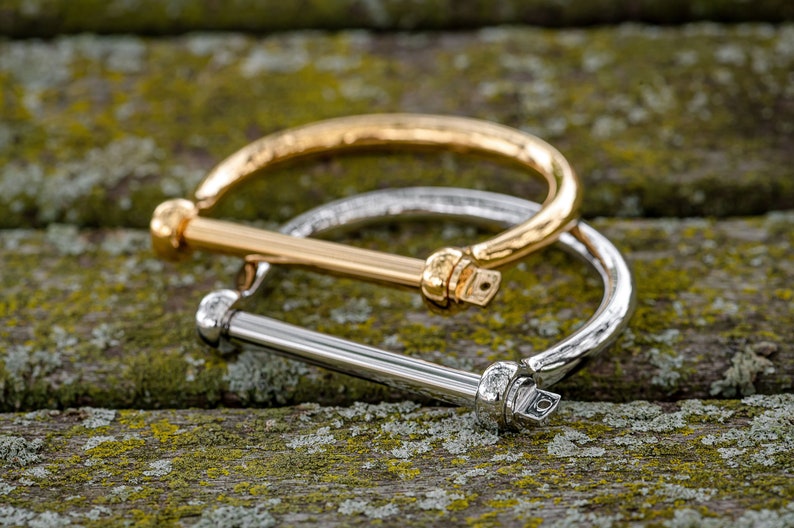 May include: Two bangle bracelets, one gold and one silver, with a textured finish. The bracelets are stacked on top of each other and have a bar closure.