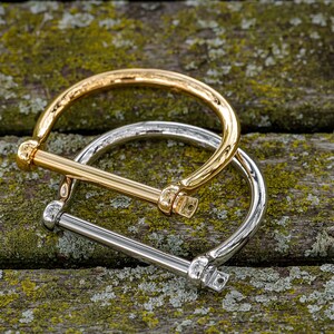 May include: Two metal bracelets, one gold and one silver, are stacked on top of each other. The bracelets are both in the shape of a circle with a straight bar across the top.