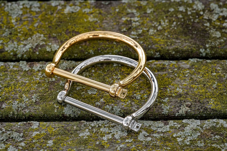 May include: Two metal bracelets, one gold and one silver, are stacked on top of each other. The bracelets are both in the shape of a circle with a straight bar across the top.
