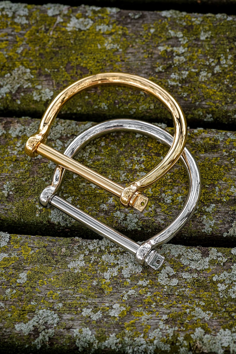 May include: Two metal bracelets, one gold and one silver, with a simple, minimalist design. The bracelets are stacked on top of each other and are lying on a wooden surface with green moss.