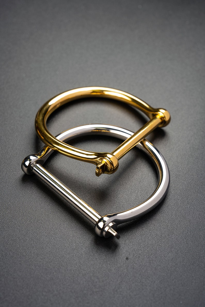 May include: Two metal bracelets, one gold and one silver, with a simple, minimalist design. The bracelets are both open and have a hinged clasp.