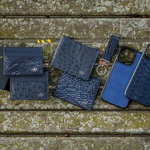 May include: A collection of blue leather goods, including wallets, phone cases, and a key chain. The items are arranged on a wooden surface and feature the A&V Nomad logo.