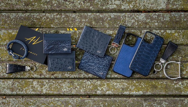 May include: A collection of blue leather goods, including wallets, phone cases, and a key chain. The items are arranged on a wooden surface and feature the A&V Nomad logo.