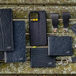 May include: A collection of black leather goods, including wallets, card holders, a phone case, and keychains. The items are arranged on a wooden surface and feature a textured, embossed design. The brand name "A/V Handmade Miami" is visible on some of the items.