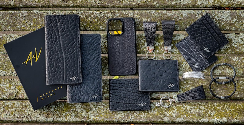 May include: A collection of black leather goods, including wallets, card holders, a phone case, and keychains. The items are arranged on a wooden surface and feature a textured, embossed design. The brand name "A/V Handmade Miami" is visible on some of the items.