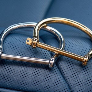May include: Two metal bracelets, one silver and one gold, with a simple bar design. The bracelets are lying on a blue leather surface.