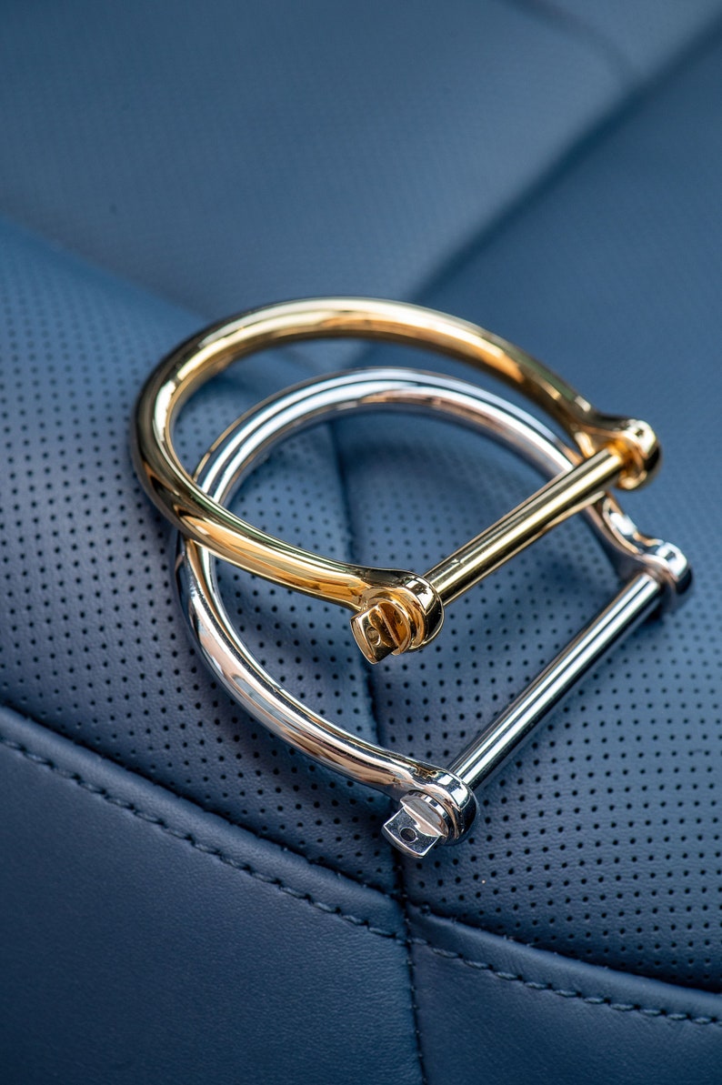 May include: Two interlocking metal bracelets, one gold and one silver, with a simple, minimalist design. The bracelets are on a blue leather surface.