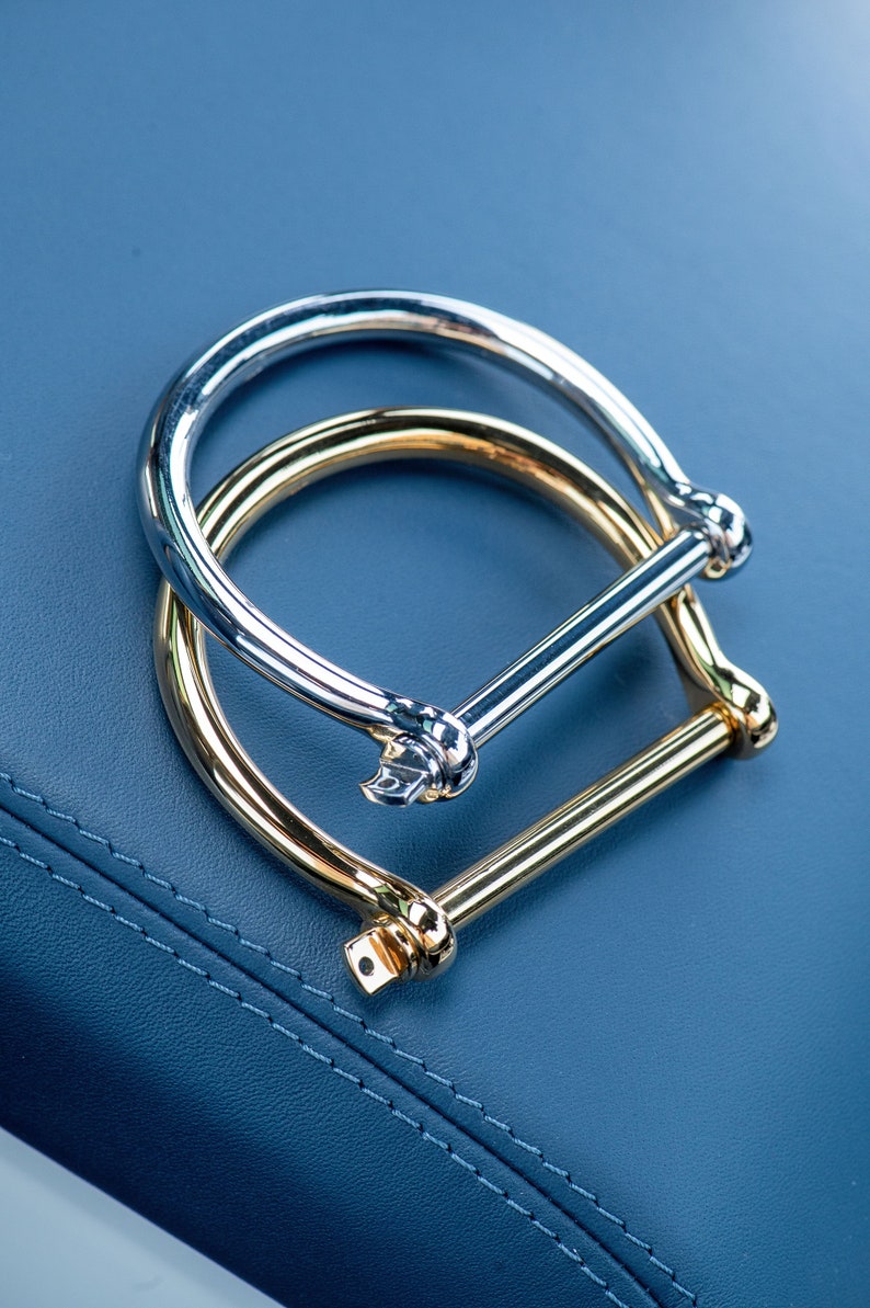 May include: Two metal bracelets, one silver and one gold, are stacked on top of each other. The bracelets are both in the shape of a circle with a straight bar across the top.