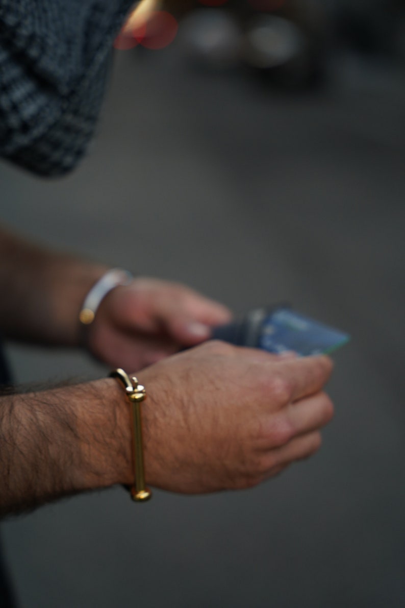 May include: A person wearing a gold bangle bracelet on their wrist. The bracelet has a simple design with two round ends.