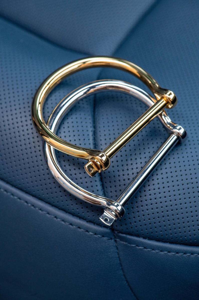May include: Two metal bracelets, one gold and one silver, with a simple, minimalist design. The bracelets are linked together by a bar that goes through both rings.