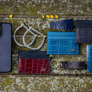 Może przedstawiać: Kolekcja sk&oacute;rzanych artykuł&oacute;w w r&oacute;żnych kolorach, w tym czarnym, niebieskim i czerwonym. Wśr&oacute;d nich znajdują się etui na telefon, portfele na karty i breloki. Wszystkie artykuły mają tłoczony wz&oacute;r sk&oacute;ry krokodyla i inicjały "HW".